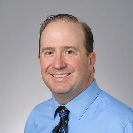 Head-and-shoulders portrait of a smiling man in a light blue dress shirt and striped tie against a gray background.